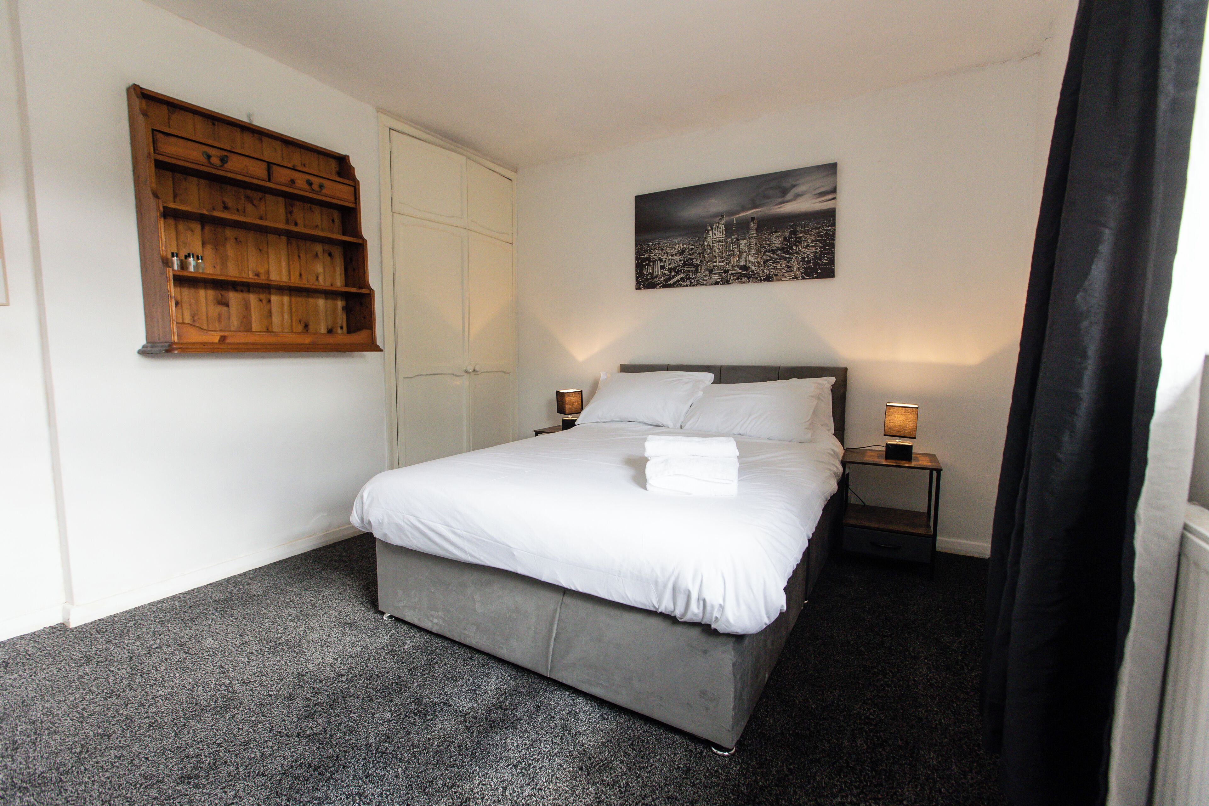 A neatly made bed with white linens, a cityscape painting on the wall, and a wooden shelf unit.