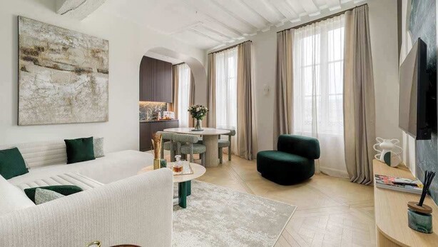 Un salon moderne avec un canapé blanc, une table basse ronde et un fauteuil vert. On y trouve également un coin repas avec une table et des chaises, et une cuisine à l’arrière-plan.