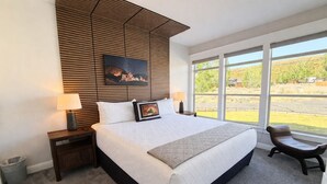 Un dormitorio con una cama grande, cabecera de madera, mesita de noche, una silla y ventana con vista a follaje.