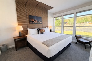 A bedroom with a large bed, a wooden headboard, a nightstand, a chair, and a window with a view of greenery.