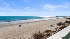Una amplia playa de arena con un muelle que se adentra en el océano, cielo despejado y algunas personas caminando.