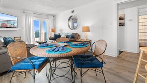 Una sala de estar moderna con una mesa redonda de mimbre, sillas con cojines azules, un sofá gris y un televisor grande en la pared.