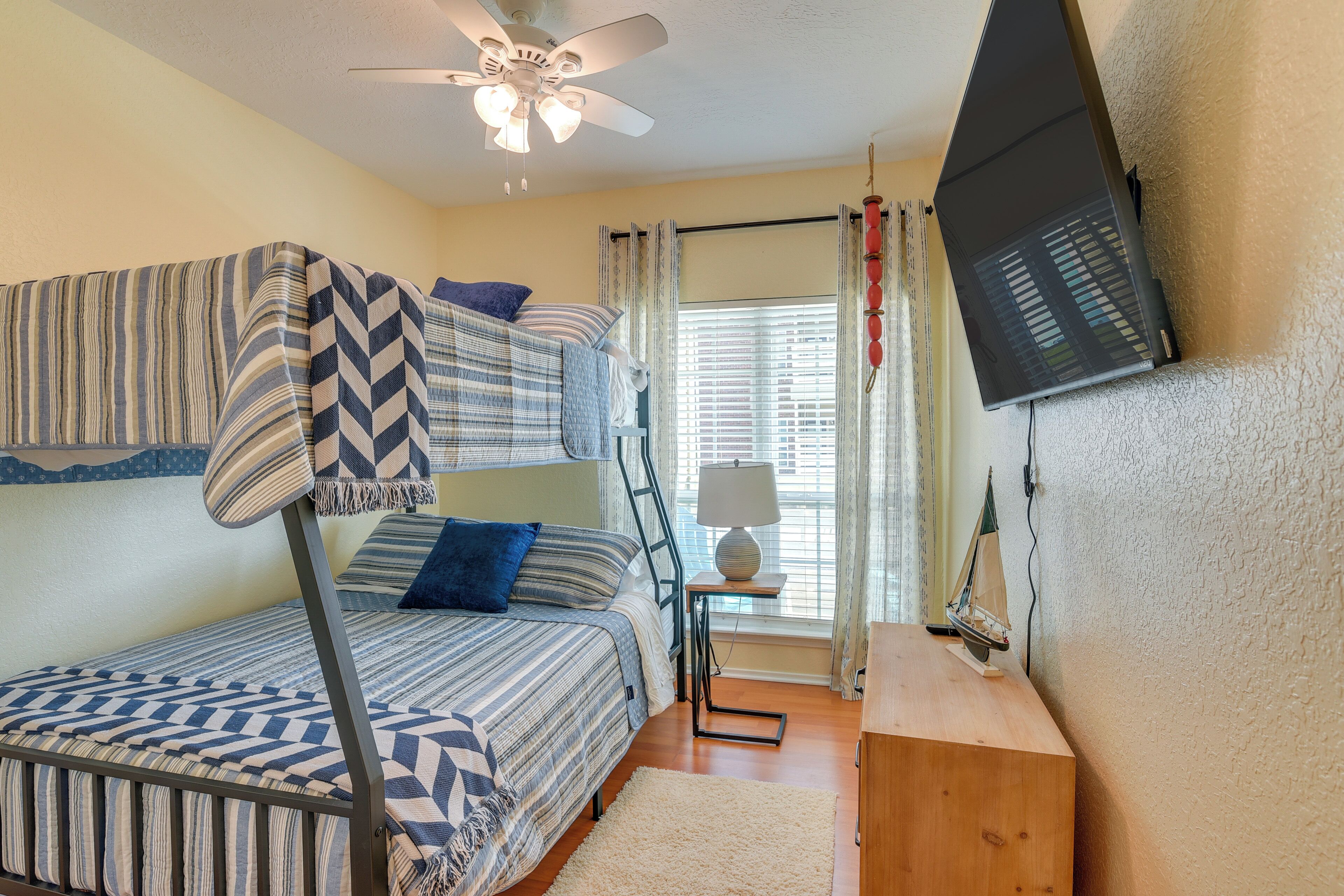 A bunk bed with patterned bedding, a ceiling fan, a small table with a lamp, and a sailboat decoration.