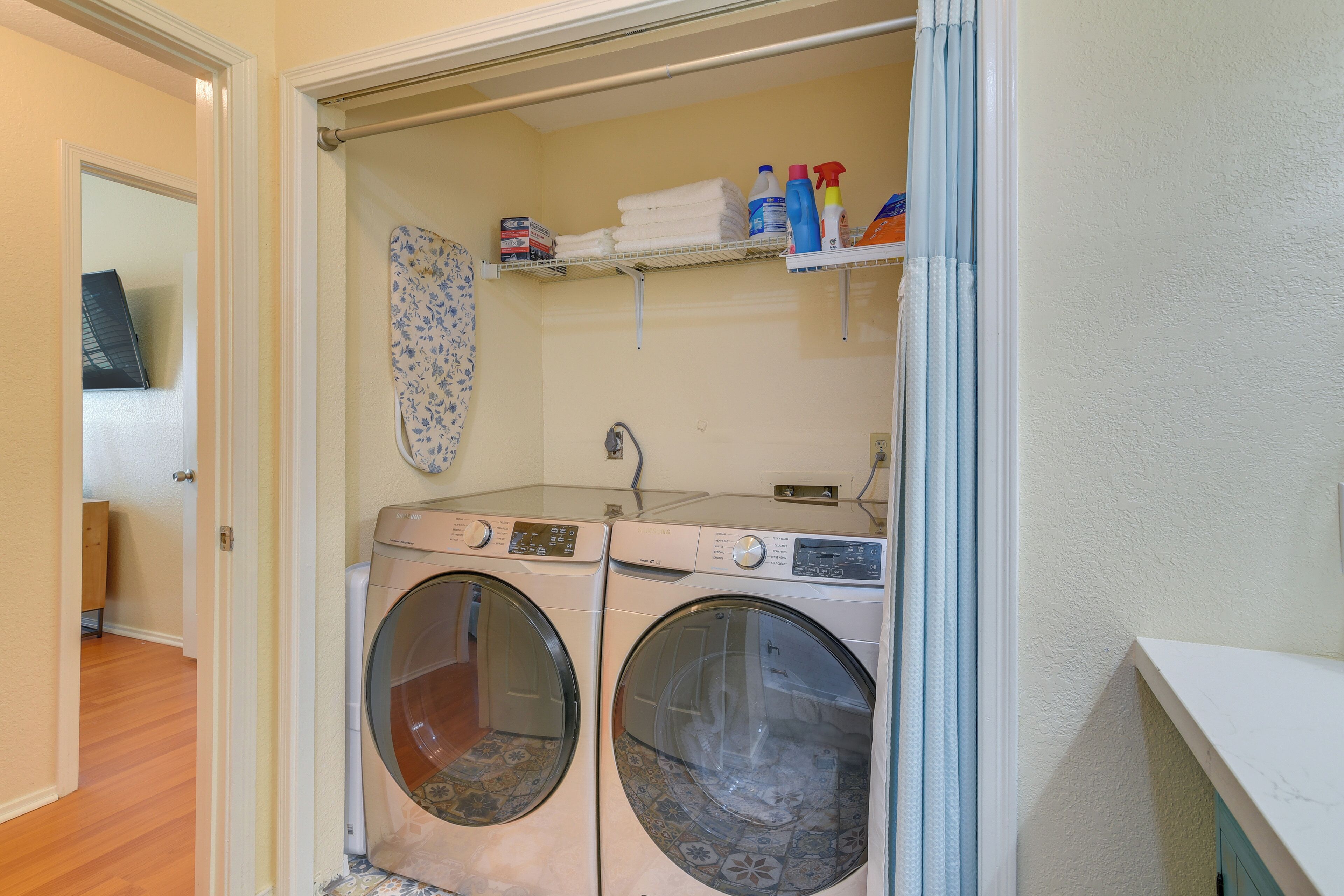 A laundry room with two front-loading washing machines, a drying rack, and shelves with laundry supplies.