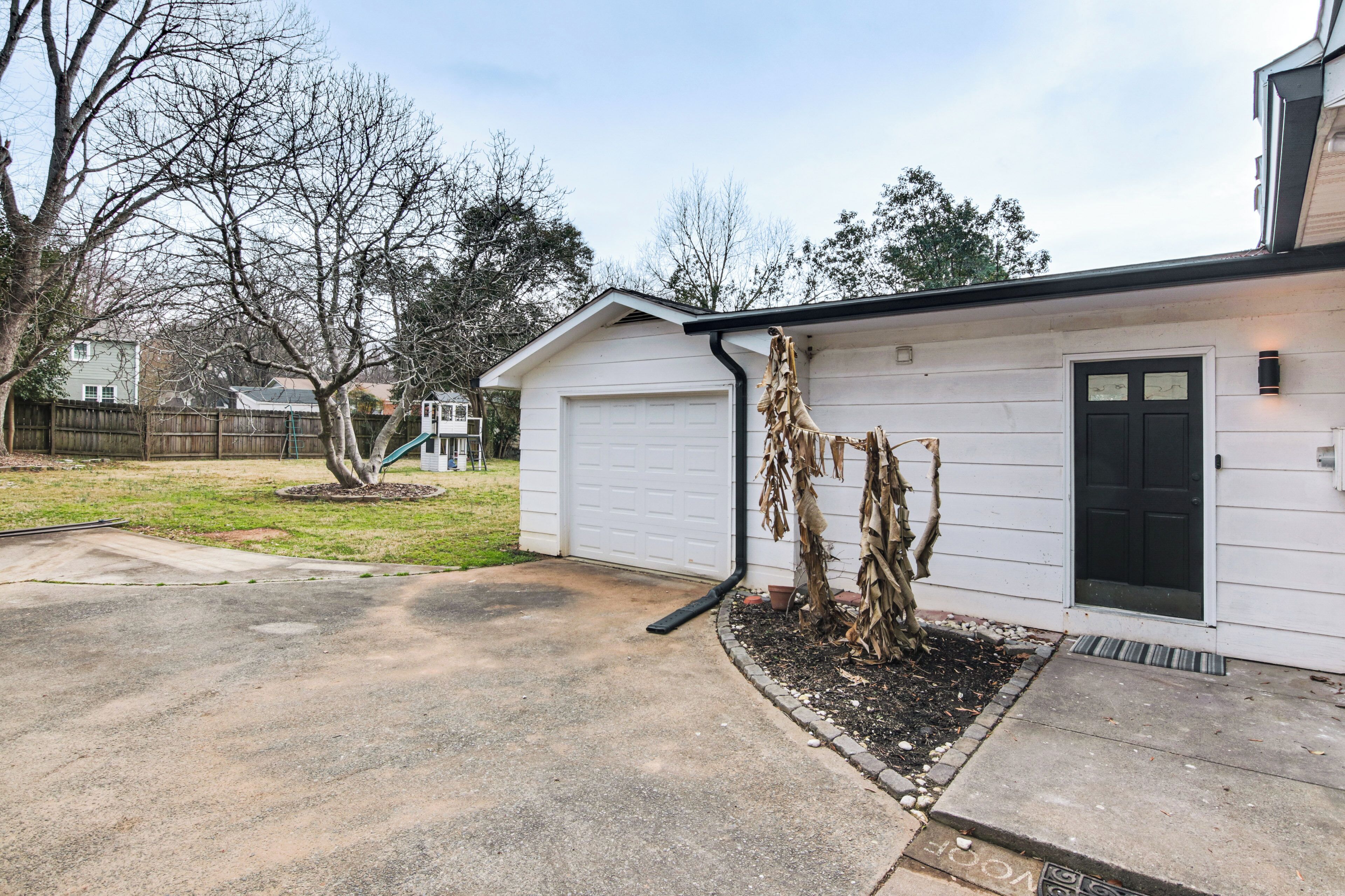 A residential garage with a white exterior, a black door, and a small porch area with a tree.