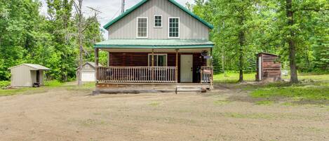 A two-story wooden cabin with a green roof, a porch, and a small shed to the left.