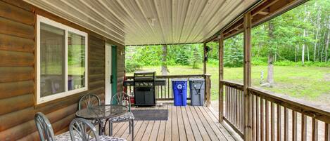 A covered deck with a wooden table and chairs, a grill, and trash bins.