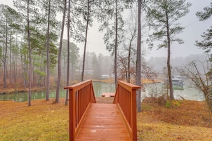 Un puente de madera que conduce a un muelle sobre aguas tranquilas, rodeado de altos pinos y una atmósfera brumosa.