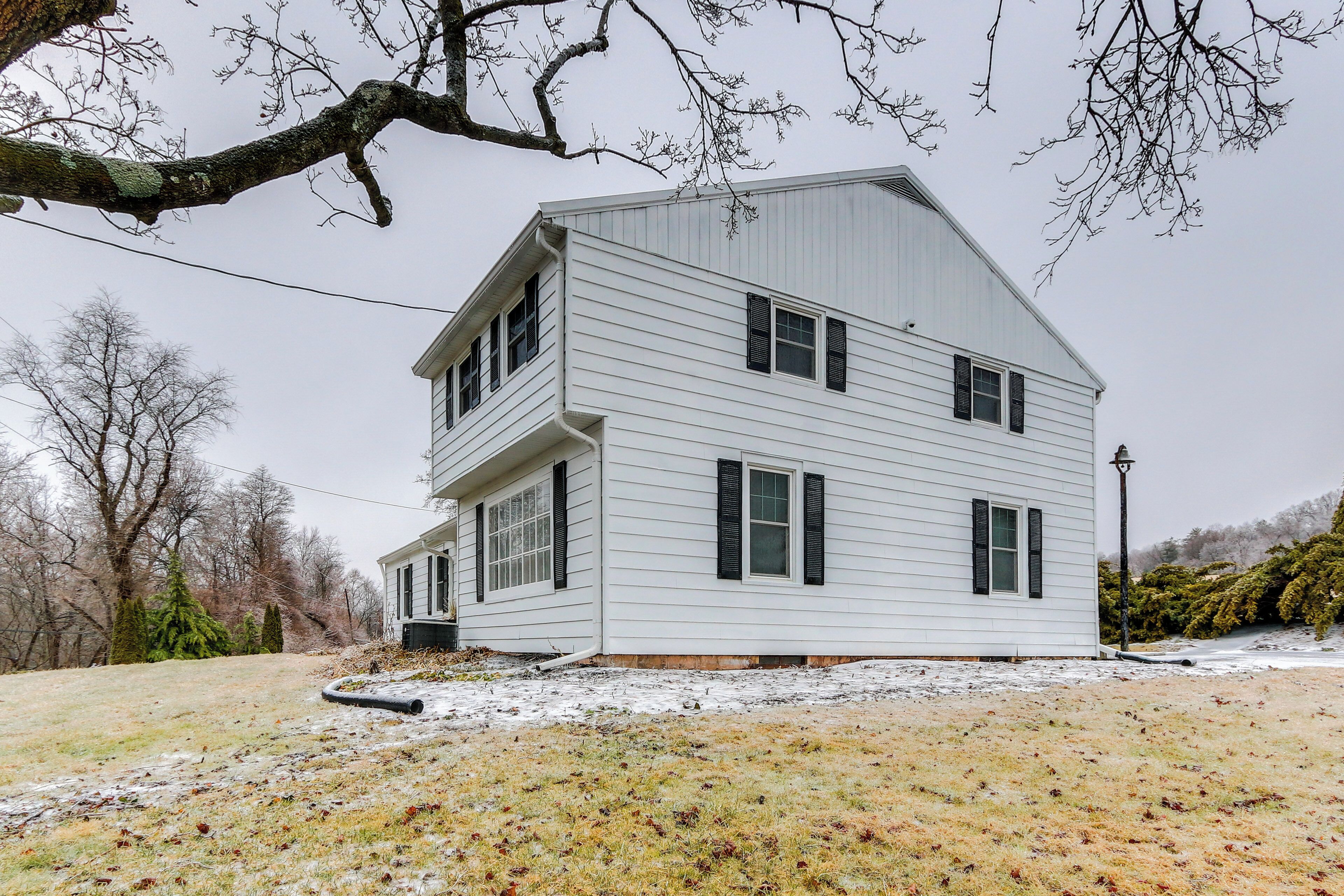 A two-story white house with black shutters, a front porch, and a yard with sparse grass and snow.