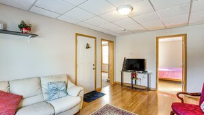 Una sala de estar con un sofá, un sillón rojo, un televisor sobre un mueble bajo y una puerta que da a un dormitorio.