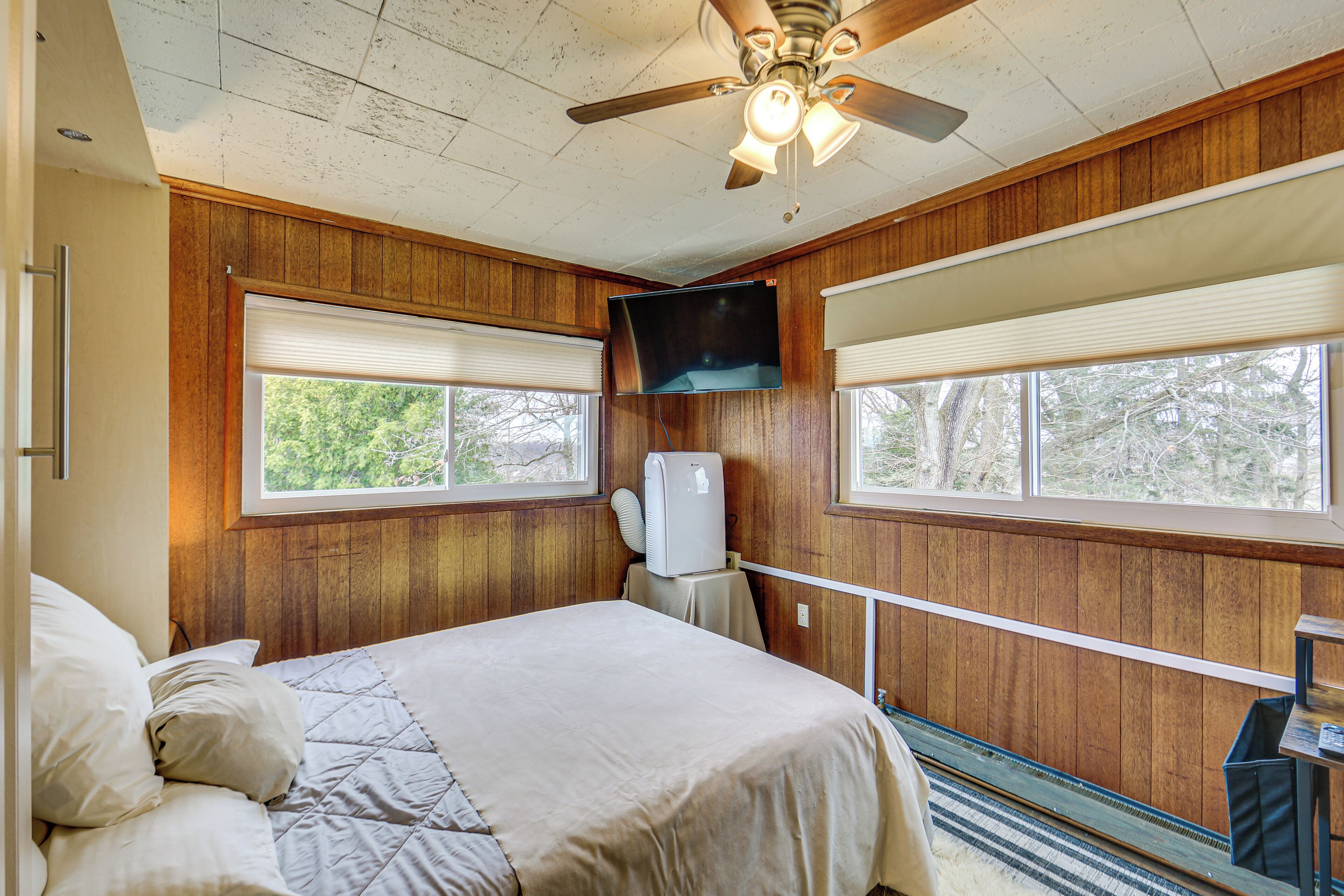 A bedroom with a bed, a ceiling fan, a TV mounted on the wall, and a window with blinds.