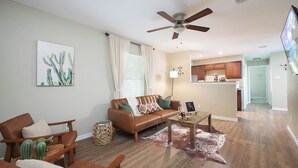 A living room with a brown leather sofa, a coffee table, and a cactus decoration. There is a ceiling fan and a kitchen area in the background.