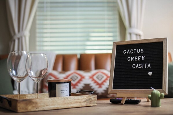 A cozy room with a wooden tray holding a candle, two wine glasses, a remote control, and a cactus on a table. A blackboard with "CACTUS CREEK CASITA" is visible.