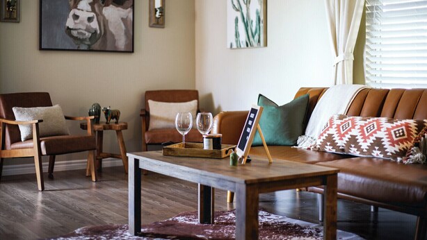 A cozy living room with a brown leather sofa, wooden coffee table, and two armchairs. There are two wine glasses on the table, a cactus painting on the wall, and a cow painting above the sofa.