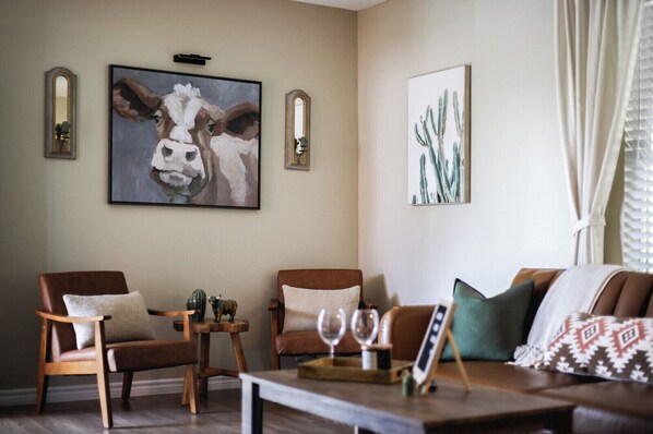 Ein Wohnzimmer mit zwei braunen Ledersesseln, einem braunen Ledersofa und einem Couchtisch mit Weingläsern. An der Wand hängen zwei gerahmte Bilder, eines von einer Kuh und eines von einem Kaktus.