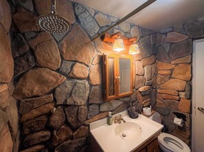 A bathroom with a stone wall, a wooden-framed mirror, a white sink, a showerhead, and a toilet. - Charming Cherokee Cabin Secluded Stay for 6 Just 10 Minutes to Tahoe! (South Lake Tahoe)