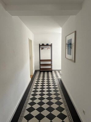 Een gang met een zwart-wit geruit vloerkleed, een kapstok met hoeden en een ingelijst schilderij aan de muur.