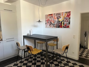 Eine kleine Küche mit Esstisch, vier Stühlen, einer Hängelampe und einem großen abstrakten Gemälde an der Wand.