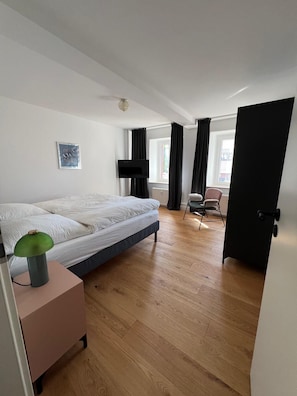 Quarto com cama, escrivaninha com luminária, cadeira, televisão e janela com cortinas.