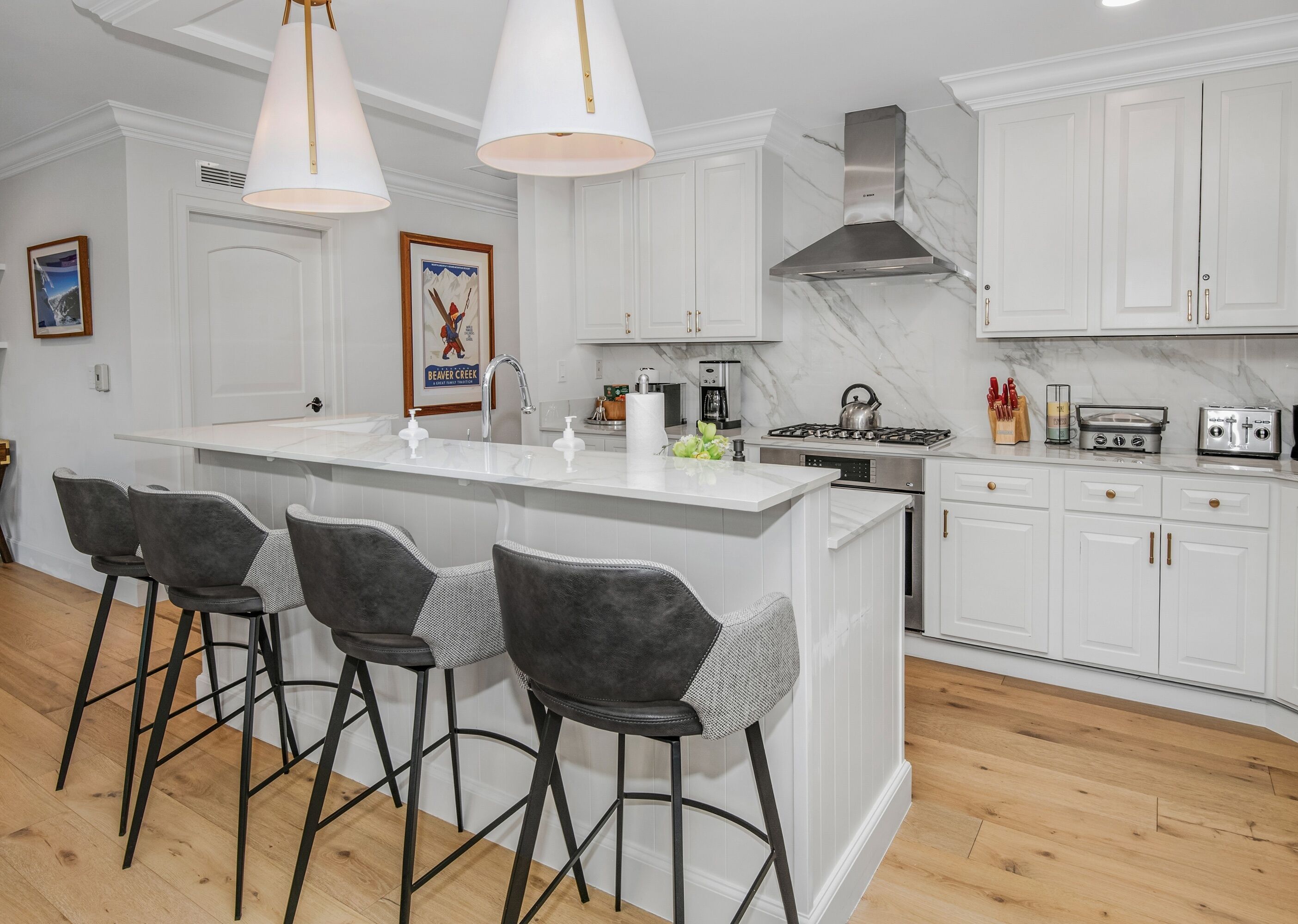 Cocina moderna con muebles blancos, una isla central con taburetes y electrodomésticos de acero inoxidable, además de iluminación colgante.