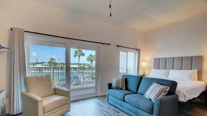 Ein Hotelzimmer mit einem Bett, einem blauen Sofa, einem weißen Sessel und einem großen Fenster mit Blick auf Palmen und einen Balkon.