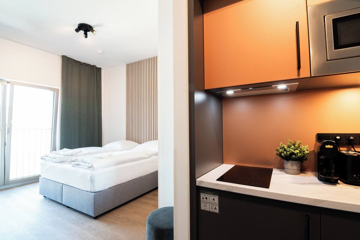 A modern hotel room with a bed, a small kitchenette, and a view of the outdoors.