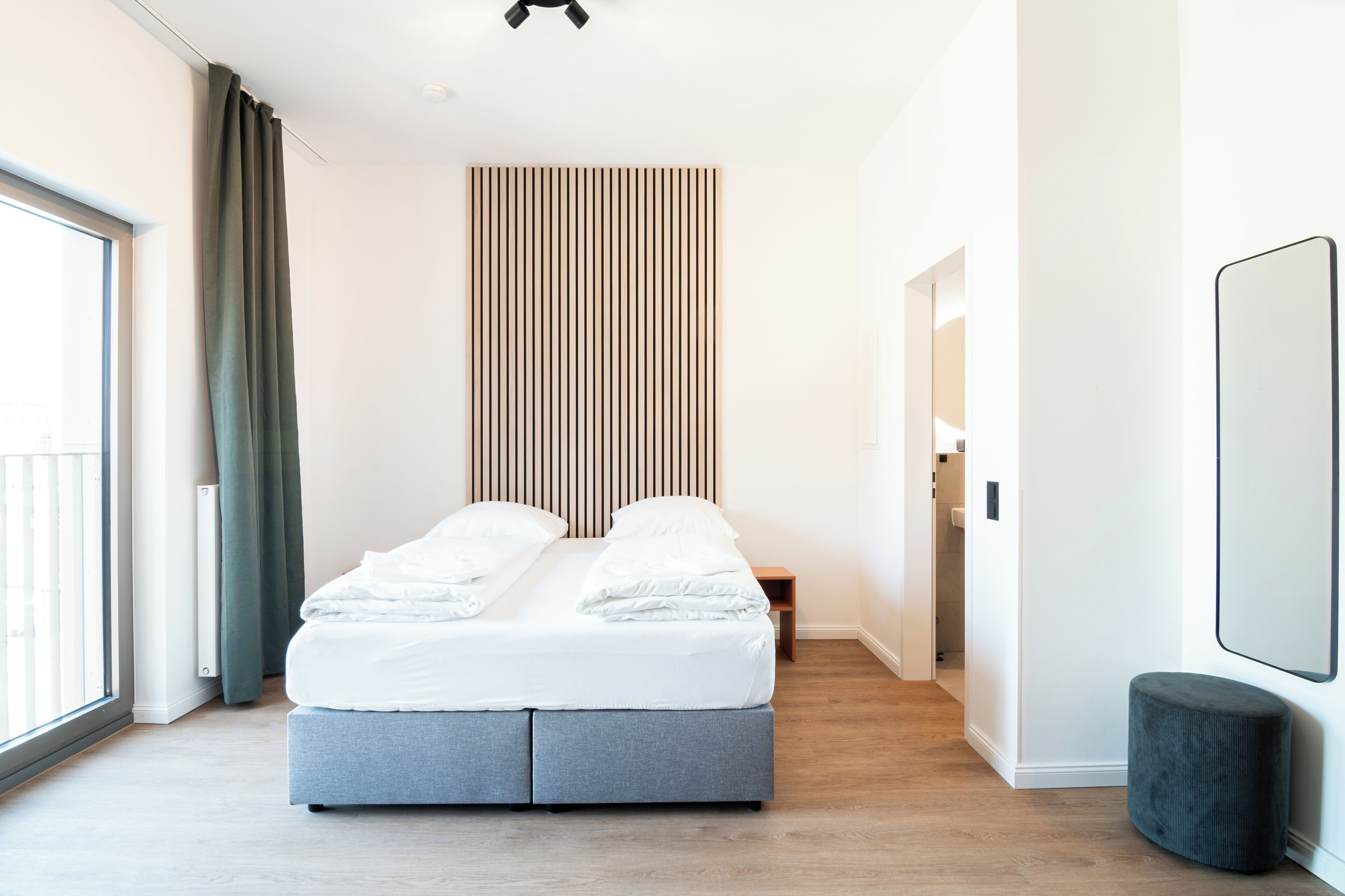 Een moderne slaapkamer met een groot bed, een houten latten hoofdboard, een nachtkastje, een klein krukje en een grote spiegel aan de muur.
