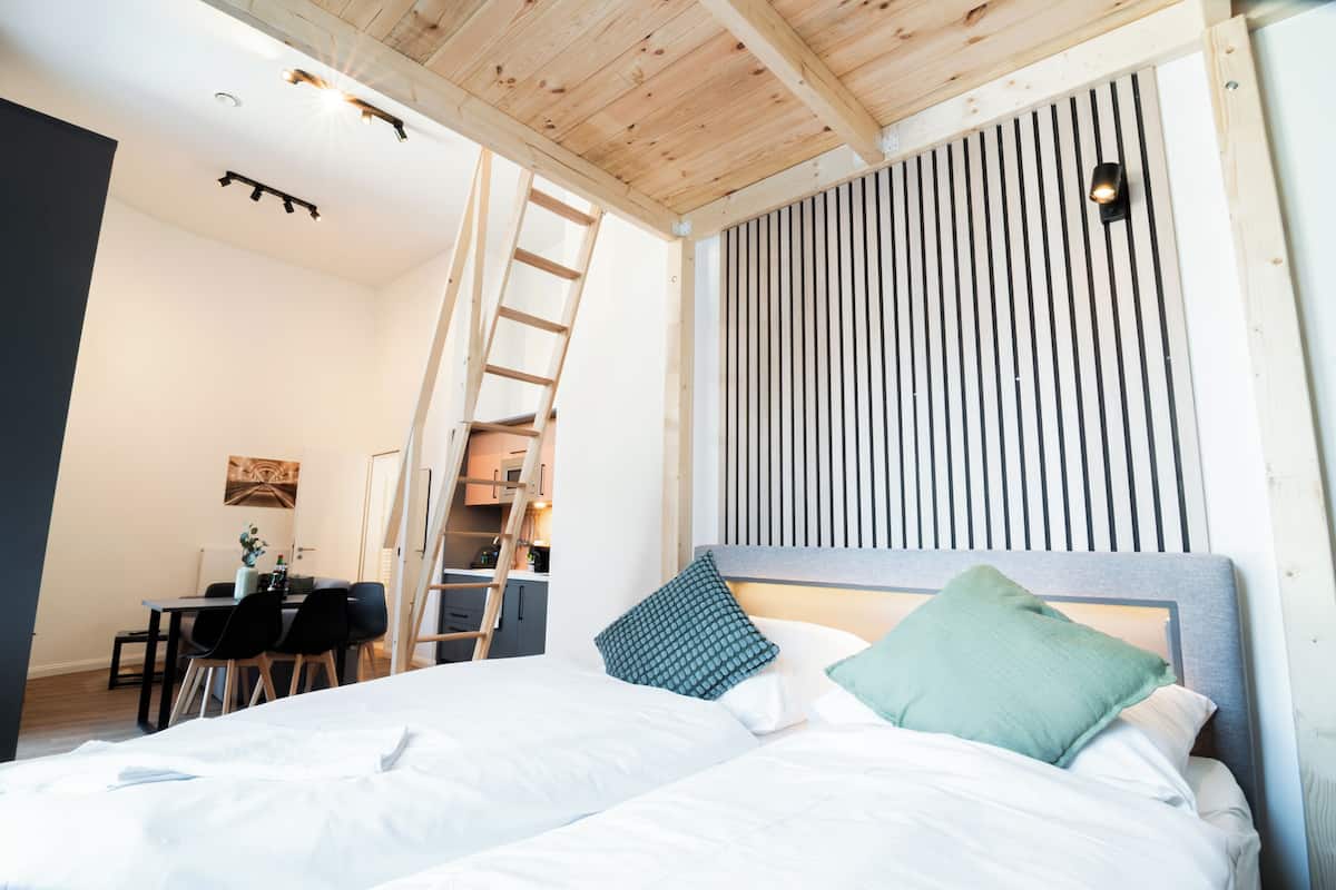 A bedroom with a bed, pillows, a desk with chairs, a ladder, and a sloped ceiling.