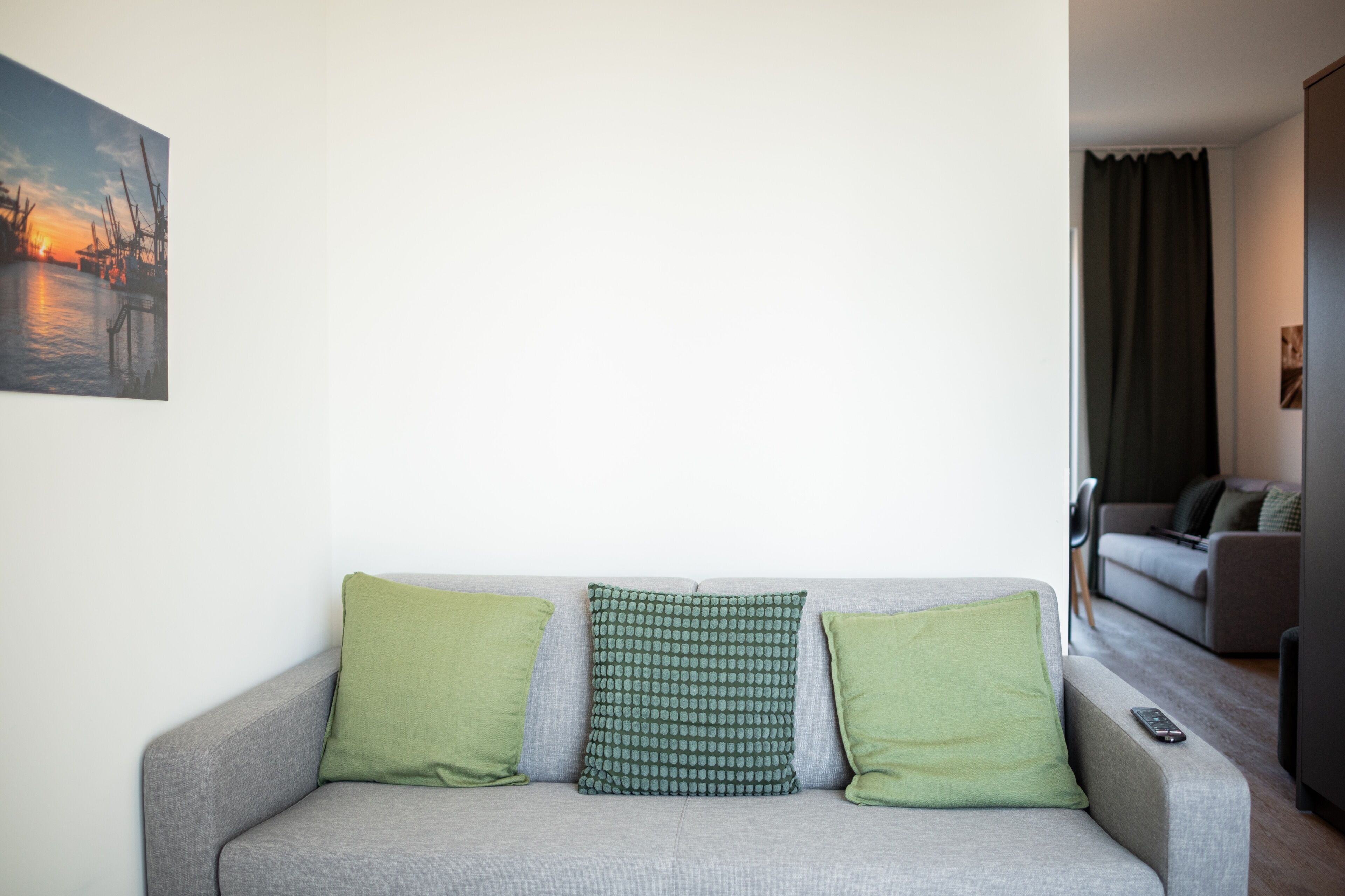 A grey sofa with three green cushions, a remote control on the right, and a painting of a sunset on the wall.