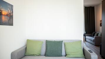 A grey sofa with three green cushions, a remote control on the right, and a painting of a sunset on the wall.