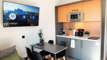 Een ruimtelijk ingerichte leefruimte met eethoek, kitchenette en een televisie aan de muur waarop een stadsgezicht te zien is.