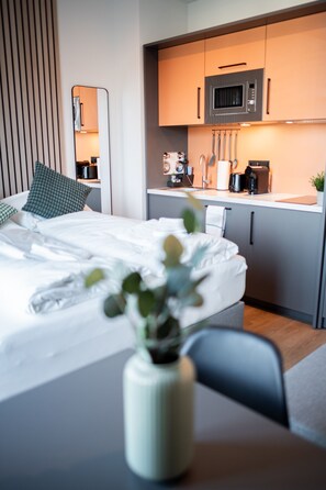 Una habitación de hotel moderna con una cama, una mesita con un florero y una kitchenette al fondo.