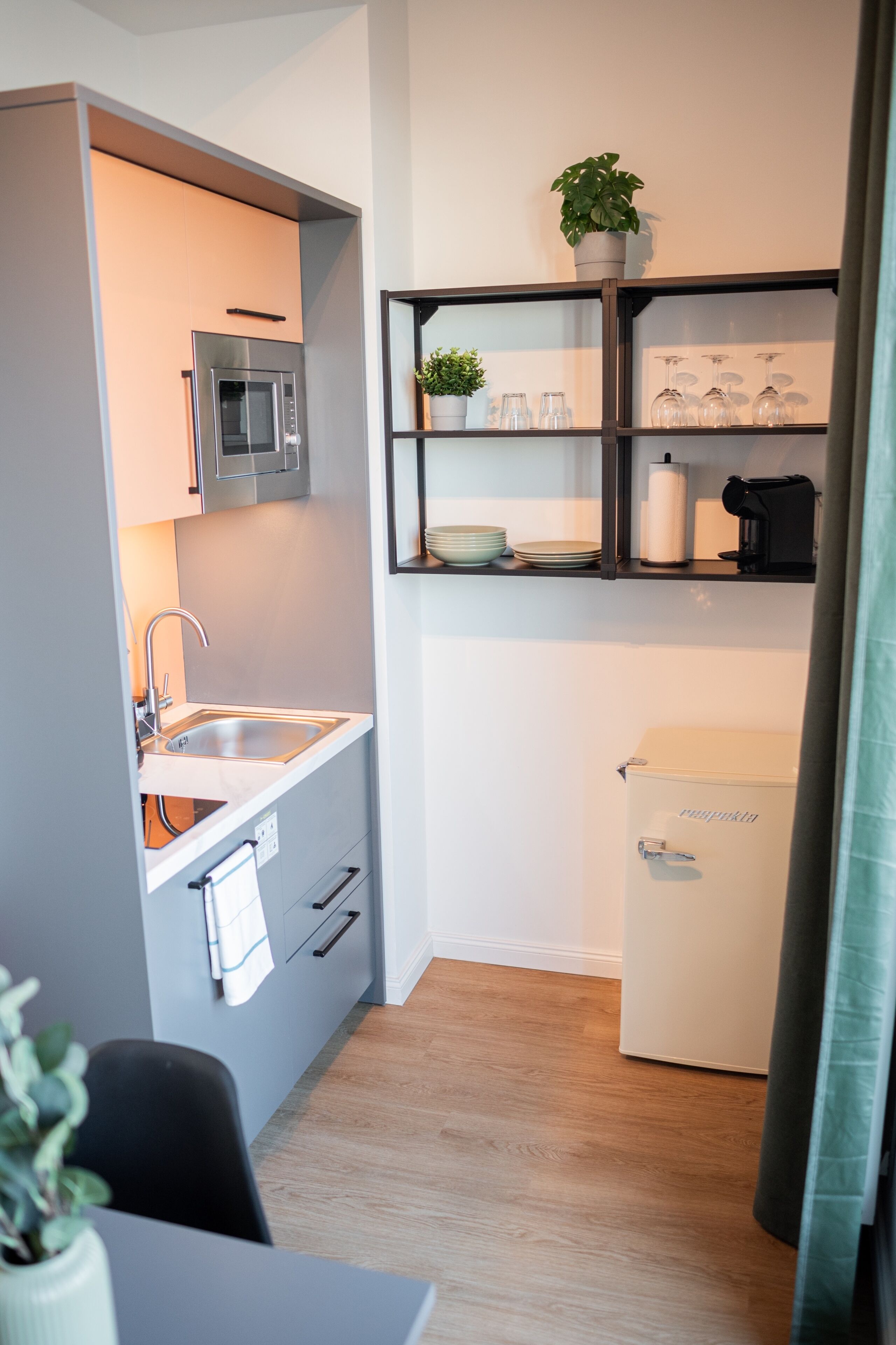 Eine kleine Küche mit integriertem Mikrowellenherd, Spüle und kompaktem Kühlschrank. Über der Spüle befindet sich ein Regal mit Gläsern und einem Blumentopf.