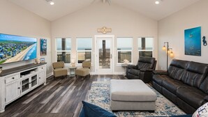 A spacious living room with a large TV, comfortable seating, a ceiling fan, and a view of the beach through large windows. - Perfectly Happy 4 Bedroom Home (Port Bolivar)