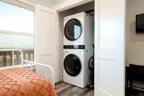 A laundry room with two washing machines, a bed with an orange patterned cover, and a view through a window. - Perfectly Happy 4 Bedroom Home (Port Bolivar)