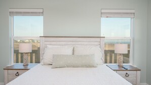 A bedroom with a large bed, two bedside tables with lamps, and a view through the windows. - Perfectly Happy 4 Bedroom Home (Port Bolivar)