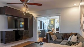 A modern living room with a flat-screen TV mounted on a dark wood cabinet, a ceiling fan, and a cozy sofa with various cushions.