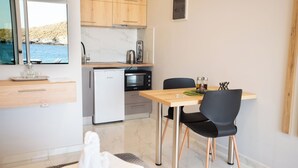 Cocina compacta con refrigerador, microondas y una pequeña isla con hervidor. Hay una mesa de comedor con dos sillas y vista al mar a través de una ventana.