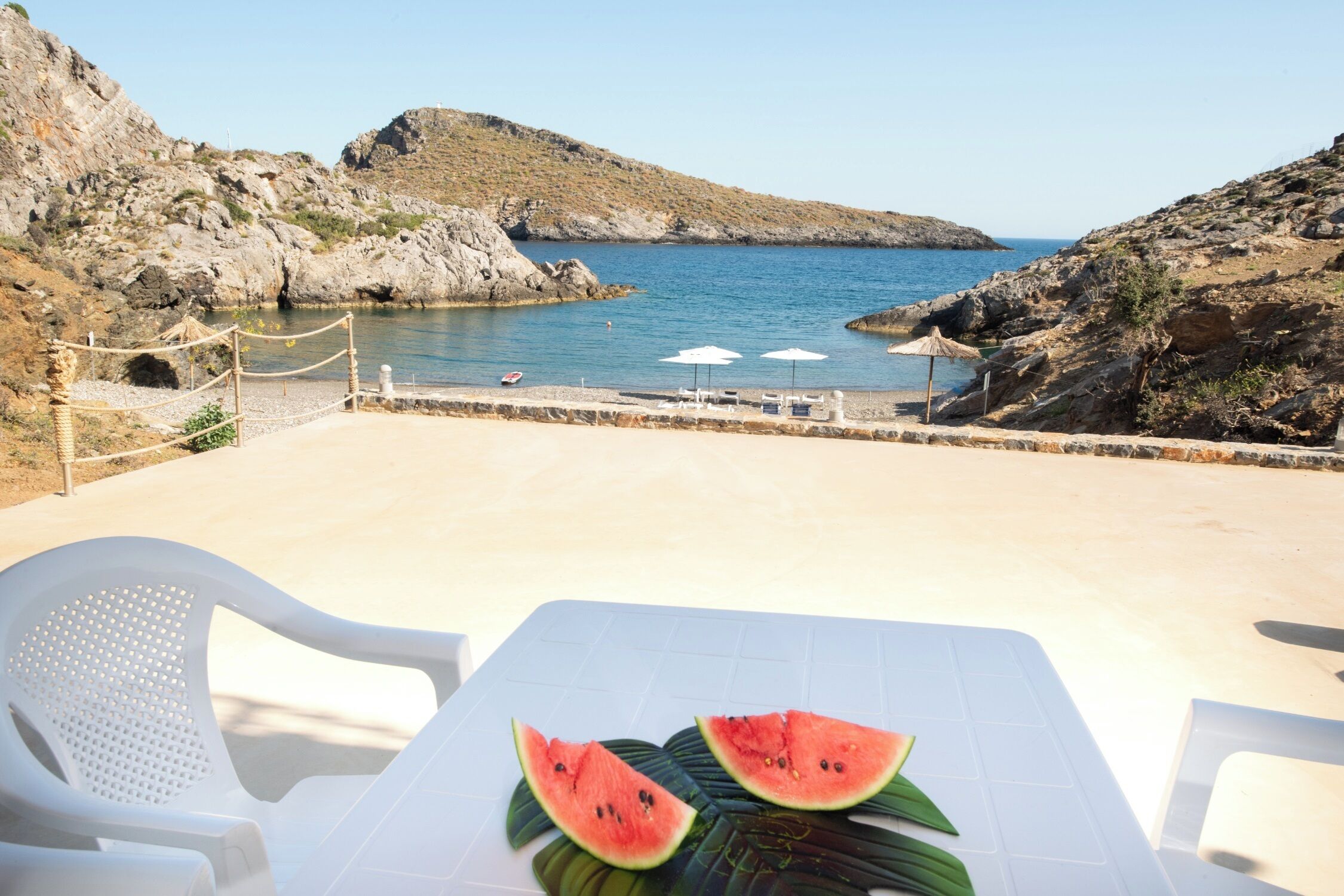 A table with a white tablecloth and two watermelon slices on it, overlooking a rocky coastline with a clear view of the sea.