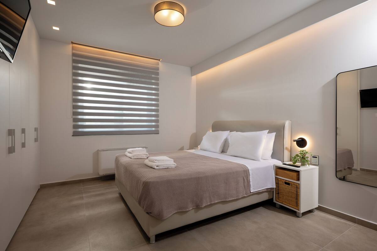 A modern bedroom with a bed, bedside tables, a mirror, and a window with blinds.