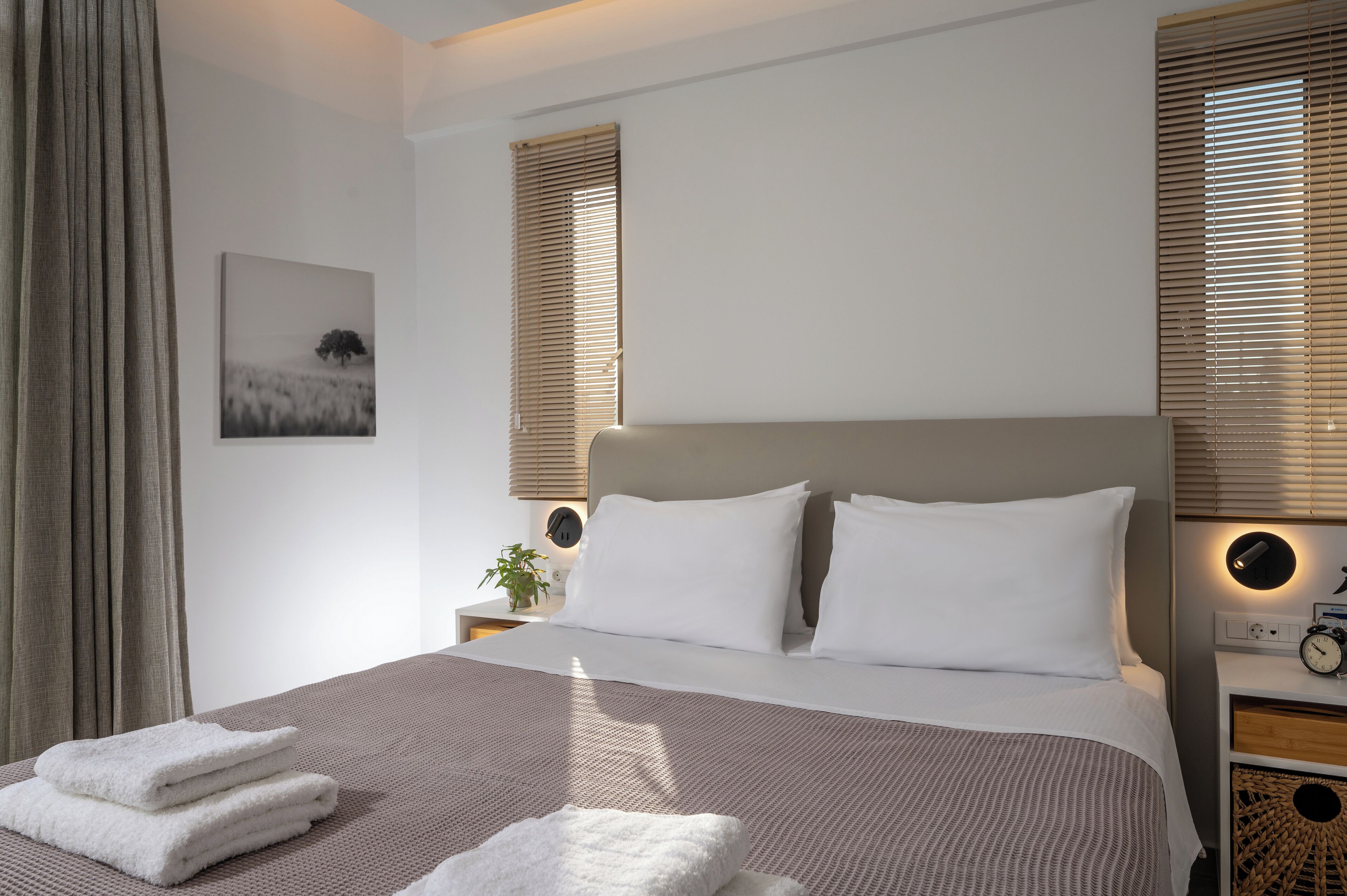 Een netjes opgemaakt bed met witte lakens, een hoofdbord en een nachtkastje met een klok en een kleine plant.