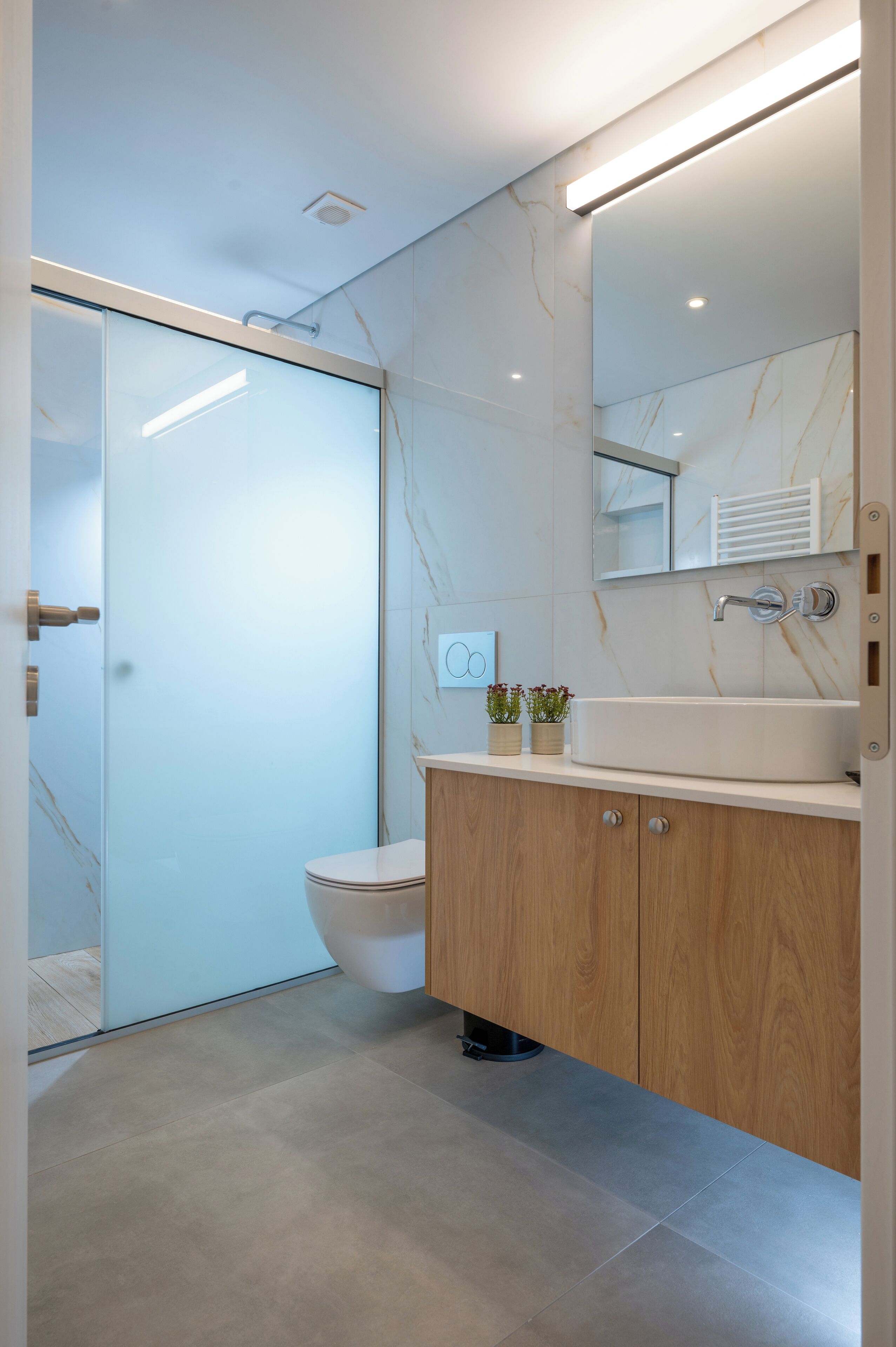A modern bathroom with a glass shower enclosure, a wooden vanity, a white sink, and a marble backsplash.