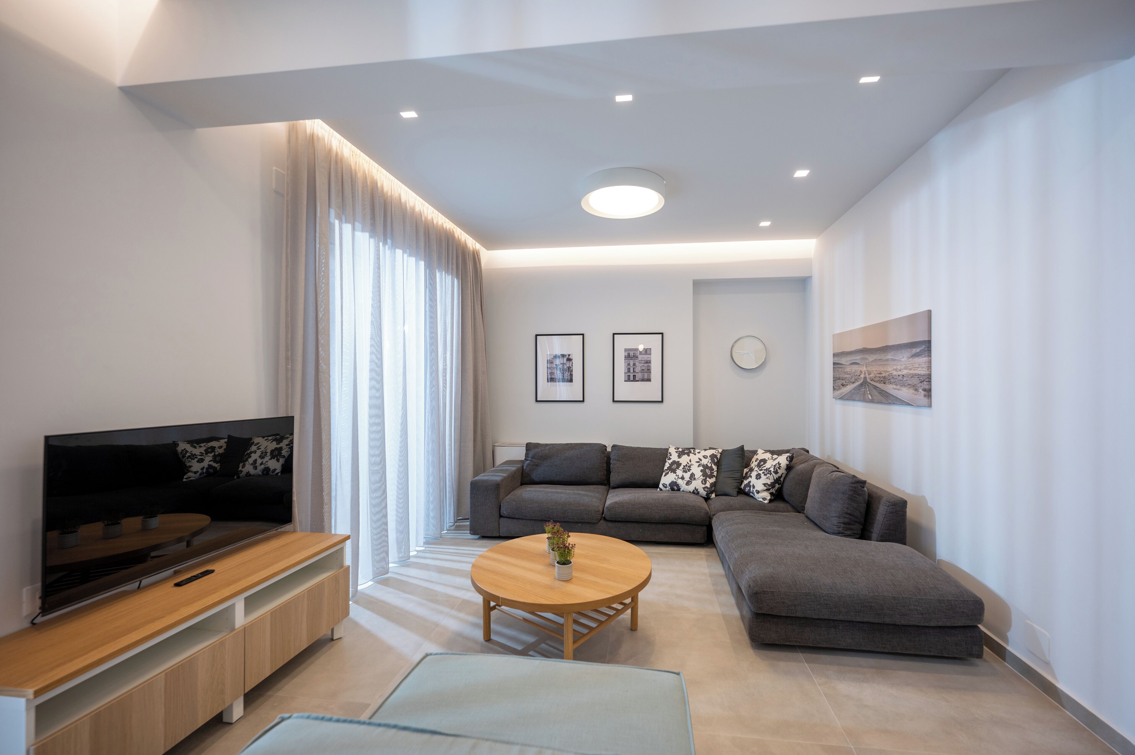 Una sala de estar moderna con un televisor de pantalla plana, sofás grises, una mesa de centro de madera y un ventanal con cortinas.