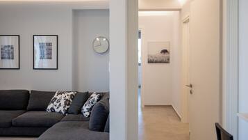 A modern living room with a dark grey sofa, two framed pictures, a wall clock, and a hallway leading to another room.