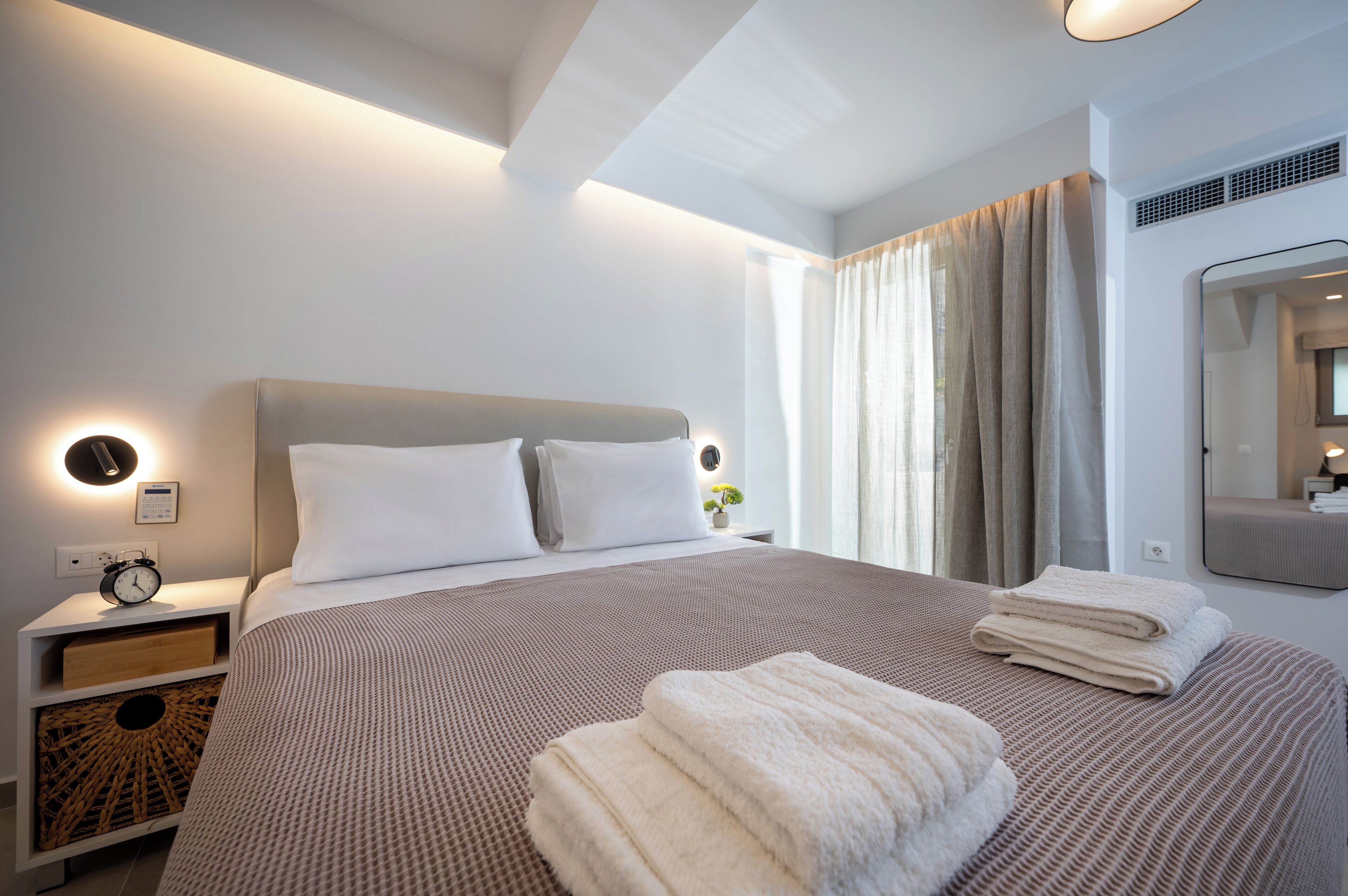 A neatly made bed with a headboard, a bedside table with a clock, and a mirror on the wall.