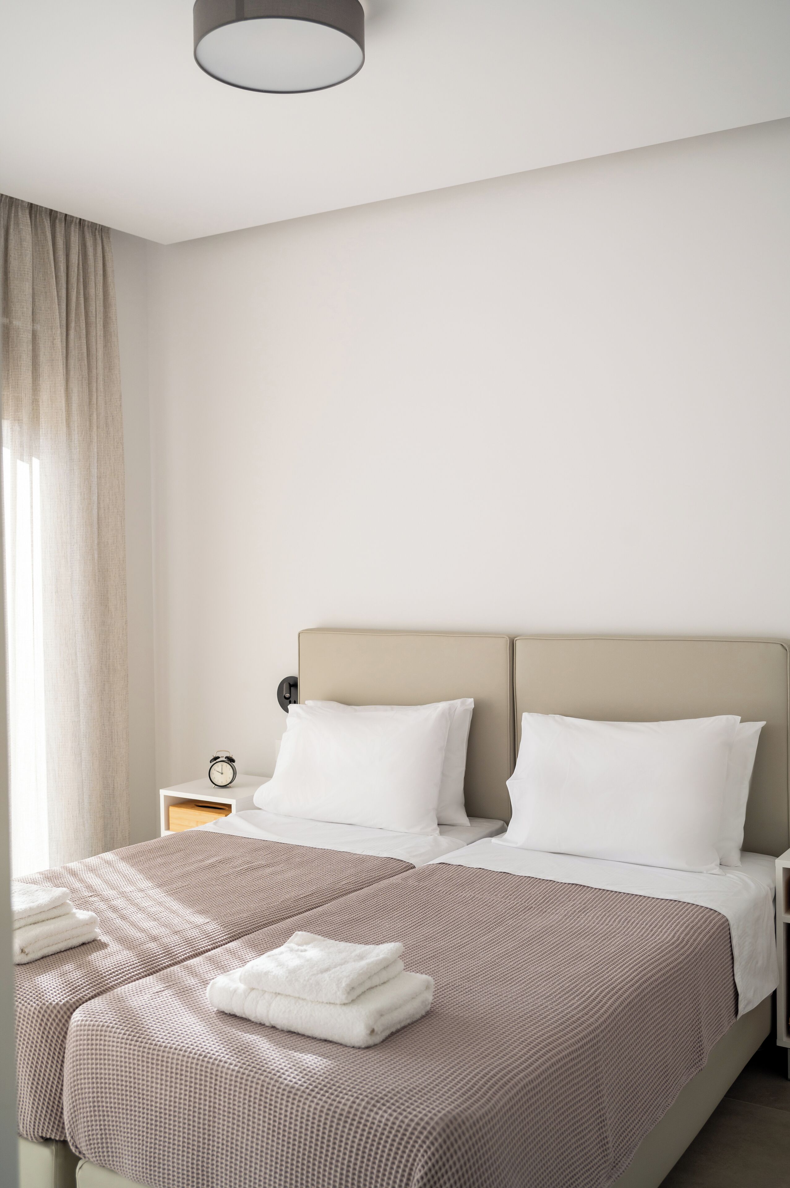Una habitación de hotel moderna con una cama grande, dos almohadas, una mesita de noche con un reloj y una ventana con cortinas.