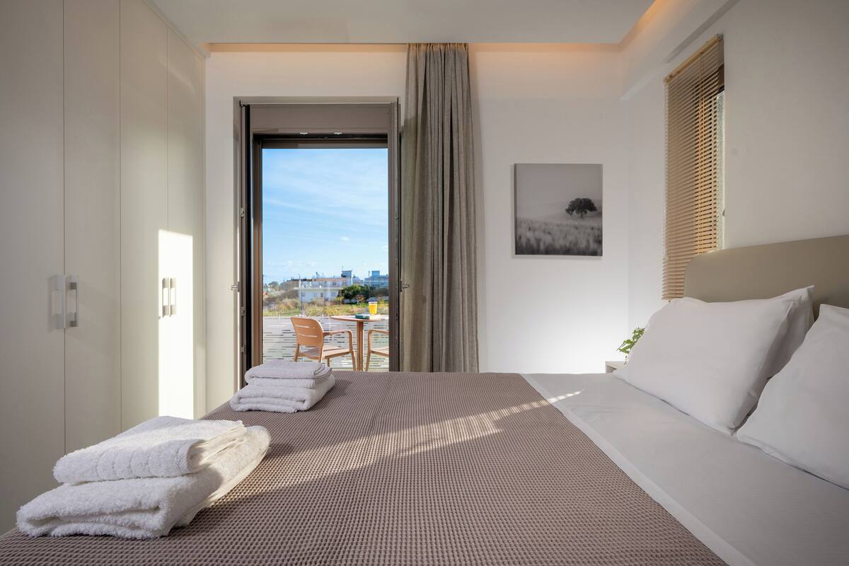 A hotel room with a large bed, a sitting area with a table and chairs, a balcony with a city view, and a window with blinds.