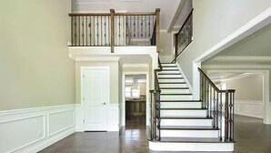 A spacious hallway with a polished wooden floor, a white door, and a staircase with a wooden railing leading to an upper level.