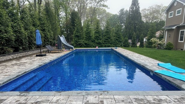 A rectangular swimming pool with a clear blue water surface, surrounded by a stone patio and a grassy area.