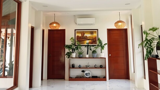 Een ruime hal met een grote geweven plafondlamp, houten deuren, een centraal geplaatst schap met planten en decoratieve objecten, en een ingelijst kunstwerk aan de muur.